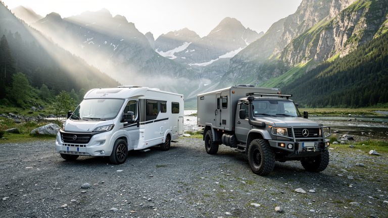 Un camping-car et un camion poids lourd aménagé stationnés côte à côte dans un paysage montagneux européen baigné par la douce lumière du matin.