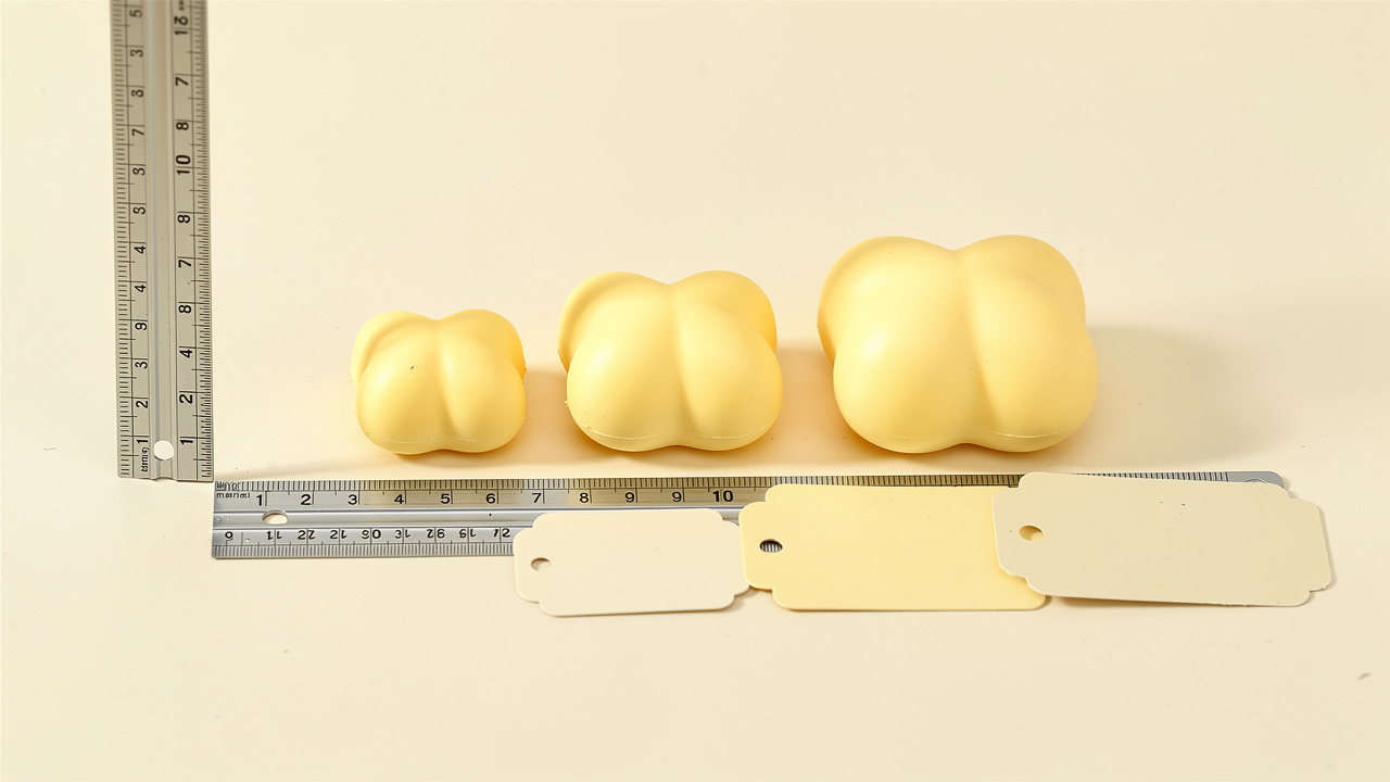 Trois squishies en forme de beurre, de petites tailles différentes, disposés à côté d’une règle non marquée et d’étiquettes neutres vierges, éclairés par une lumière naturelle.